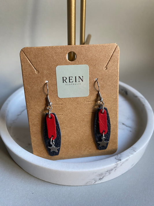 Pair of earrings (Red rounded rectangle with a silver star hanging below against a navy leather background, handing from a stainless steel hook) on a REIN branded card with a marble surface background