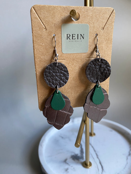 Pair of earrings (Dark Bronze circle with A medal bronze leaf shape and a green teardrop overlaid suspended below, set on a hook), on a REIN branded card with a marble surface in the background