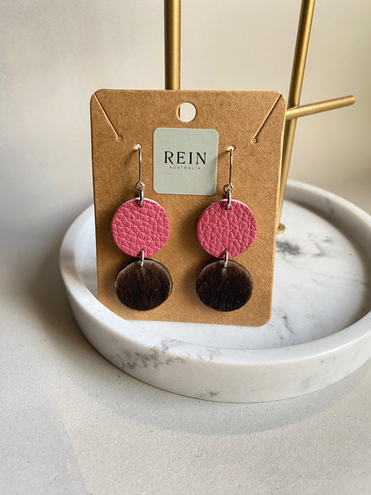 Pair of leather earrings - dark pink pebble leather circle with a dark brown hair-on hide circle suspended below, with a silver hook against a marble, grey and gold background with REIN branding