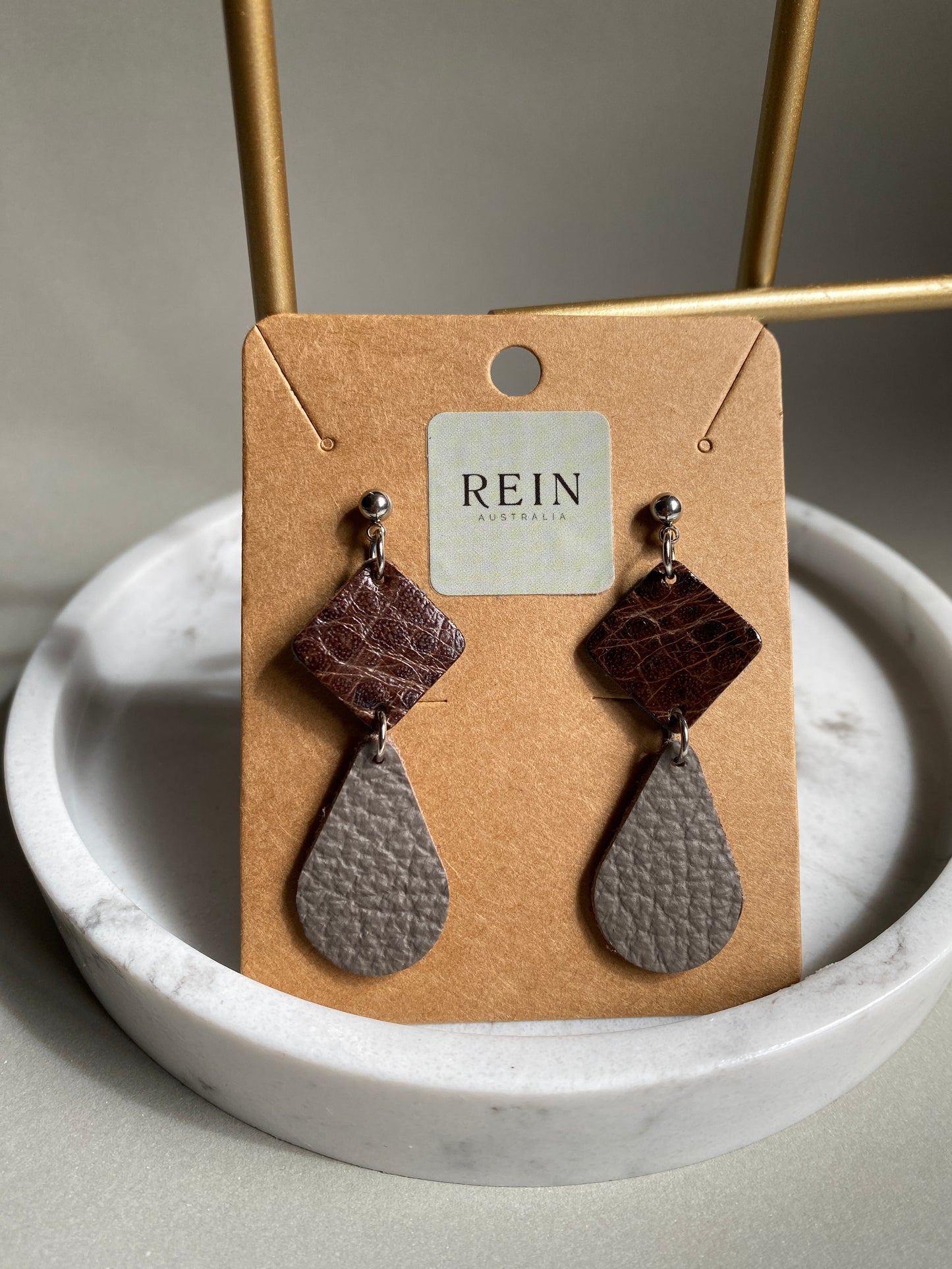 Pair of leather earrings which have a dark brown square with a grey teardrop suspended below, on a REIN branded card against a neutral background