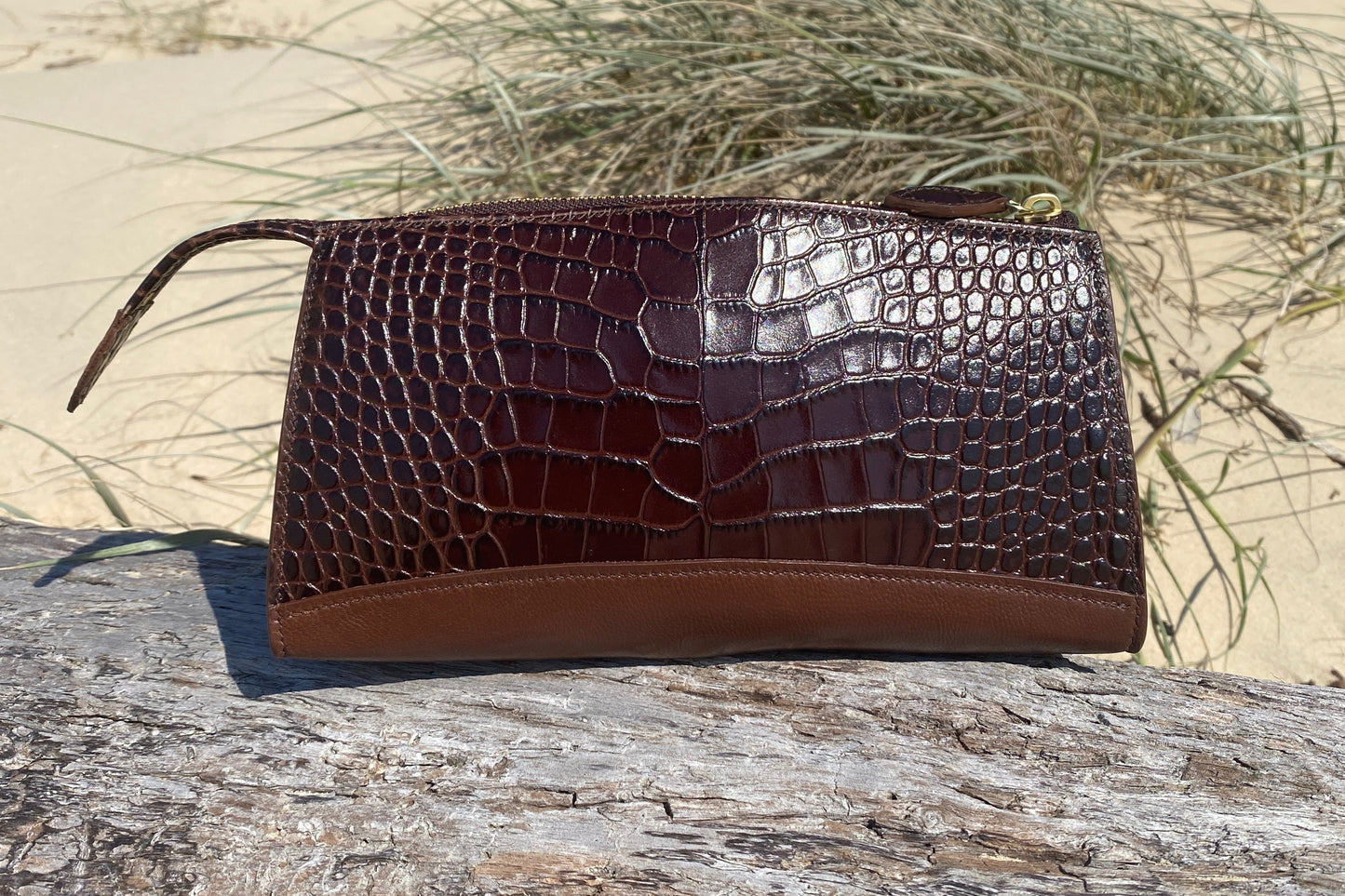 Brown crocodile-patterned clutch bag on a wooden log with 'REIN Australia' branding.