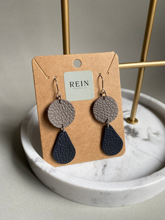 Pair of leather earrings which have a grey circle with a navy teardrop suspended below, on a REIN branded card against a neutral background