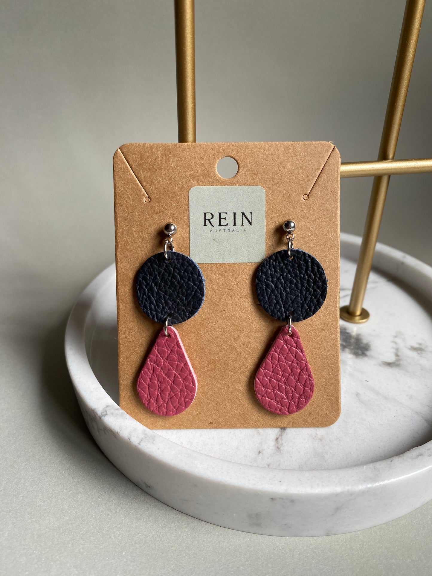 Pair of leather earrings which have a navy circle with a dark pink teardrop suspended below, on a REIN branded card against a neutral background