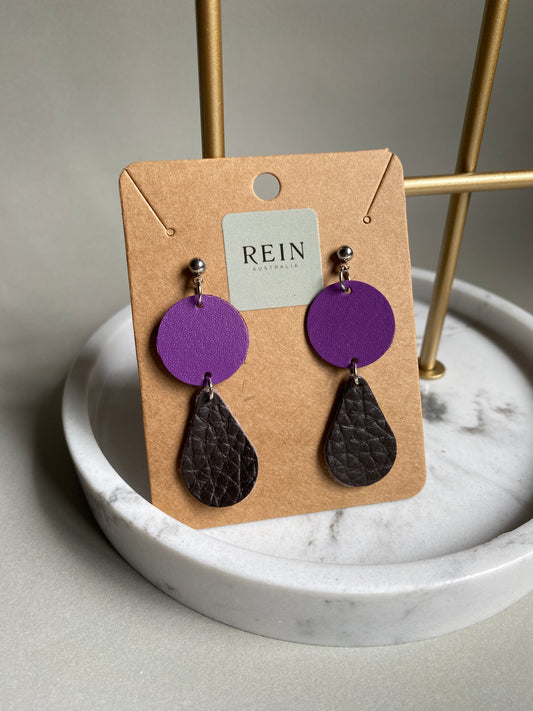 Pair of leather earrings which have a purple circle with a dark bronze teardrop suspended below, on a REIN branded card against a neutral background