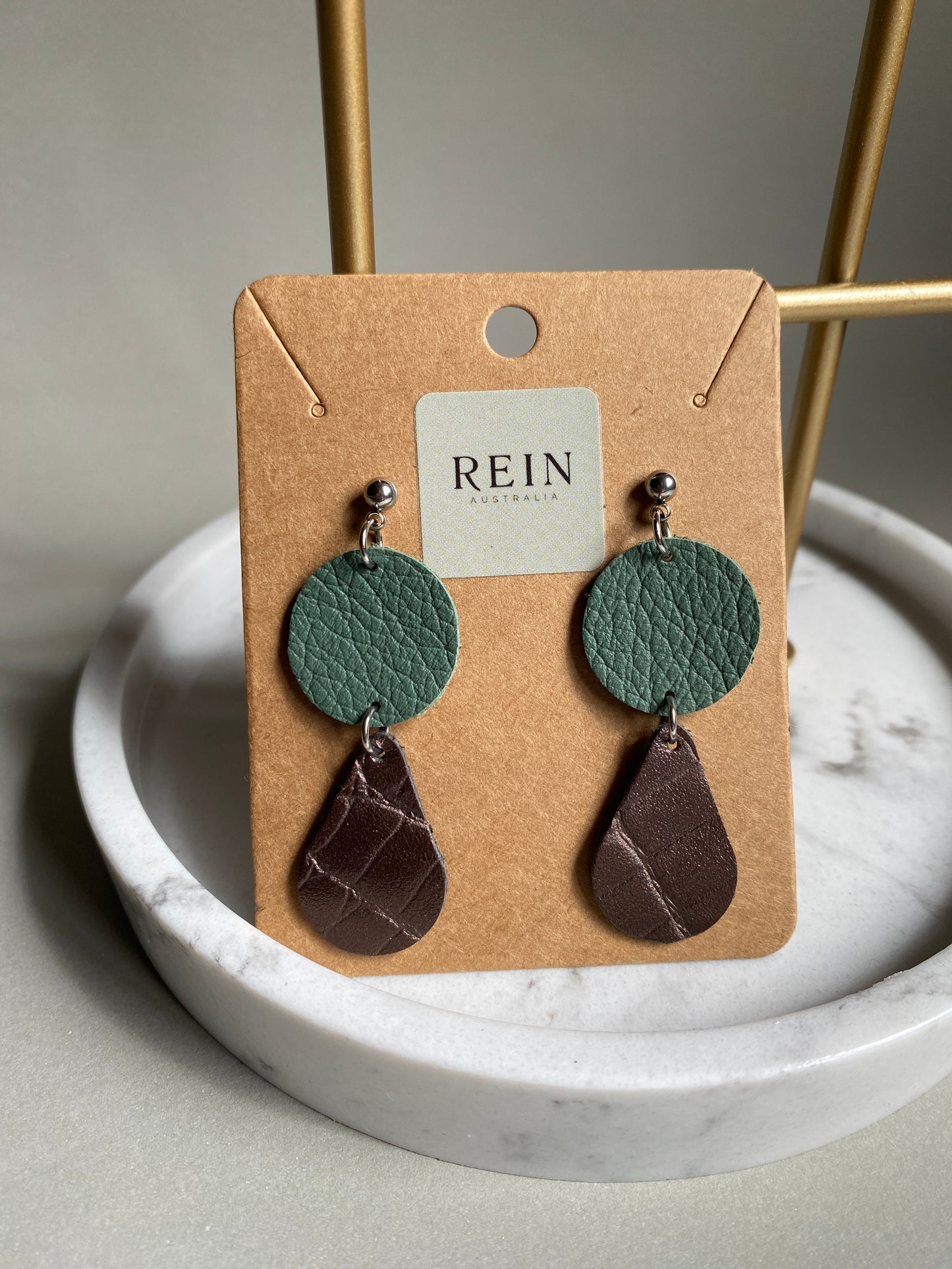 Pair of leather earrings which have a sage green circle with a medal bronze mock croc embossed leather teardrop suspended below, on a REIN branded card against a neutral background