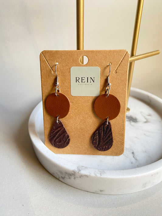 Pair of leather earrings - tan circle with a dark brown embossed teardrop suspended below, with a silver hook against a marble, grey and gold background with REIN branding