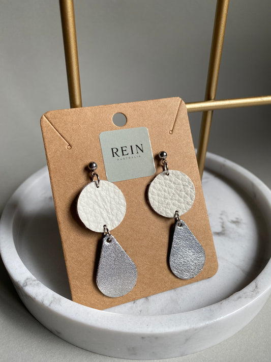 Pair of leather earrings which have a white circle with a metallic silver teardrop suspended below, on a REIN branded card against a neutral background