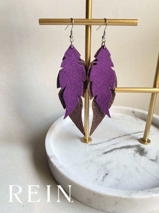 Pair of leather earrings - purple suede feather layered over a larger dark brown embossed leather feather with a silver hook against a marble, grey and gold background with REIN branding
