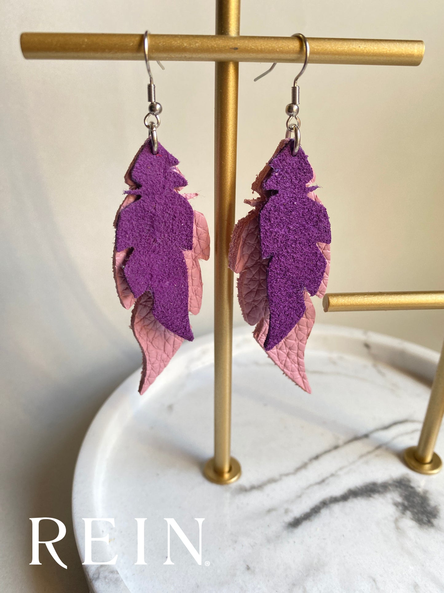 Purple and pink feather-shaped earrings on a gold stand with 'REIN' branding.
