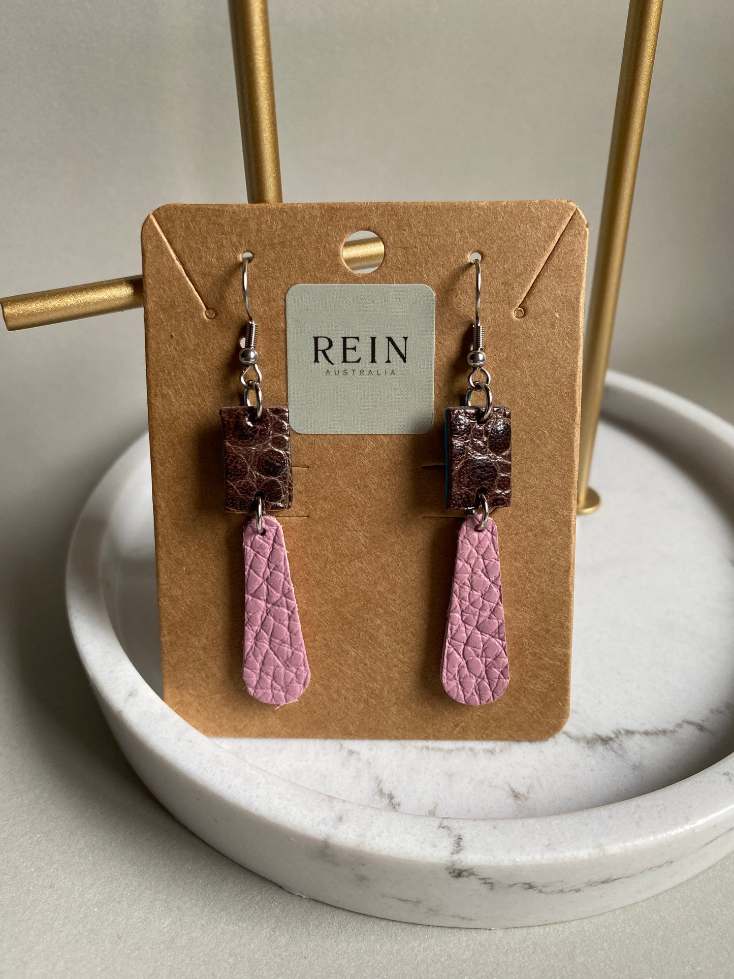 Pair of earrings - Brown Cane Toad Skin leather Rectangle with a pale pink pebble grain leather elongated teardrop shape suspended below. Earrings feature stainless steel hook and hardware. Presented on a card with REIN branding with a marble and grey background.