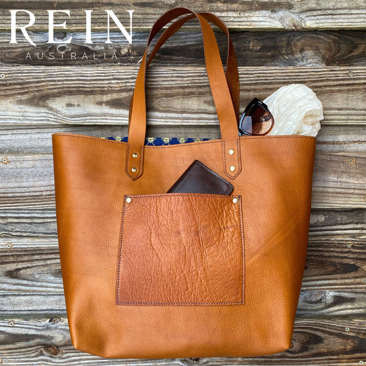 A brown leather tote bag with navy and gold-foil honey bee lining, displayed against a wooden background, with riveted straps and a front pocket visible.