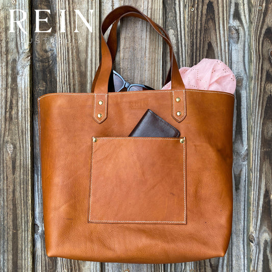 Side view of tan leather tote bag with sunglasses, scarf and a leather wallet peeping out, with a wooden background.