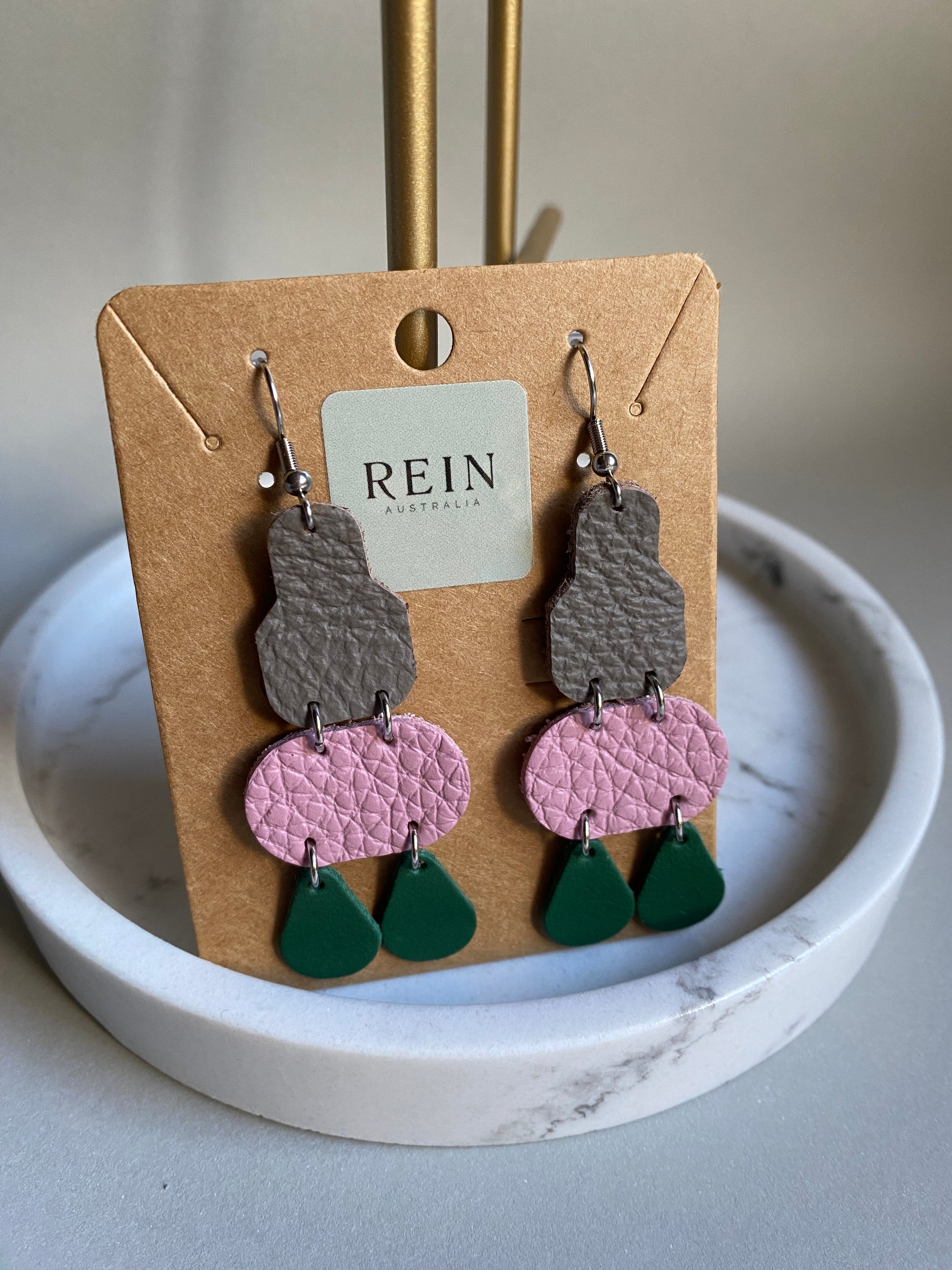 Colorful earrings on a REIN branded card with a marble surface background
Earrings are a grey leather irregular shape with a pink leather oval suspended below, with two green teardrops suspended from the pink oval. 