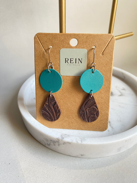 Pair of leather earrings - aqua circle with a dark brown embossed teardrop suspended below, with a silver hook against a marble, grey and gold background with REIN branding
