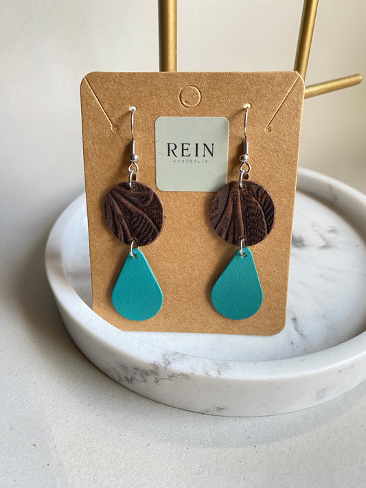 Pair of leather earrings - dark brown embossed circle with an aqua teardrop suspended below, with a silver hook against a marble, grey and gold background with REIN branding