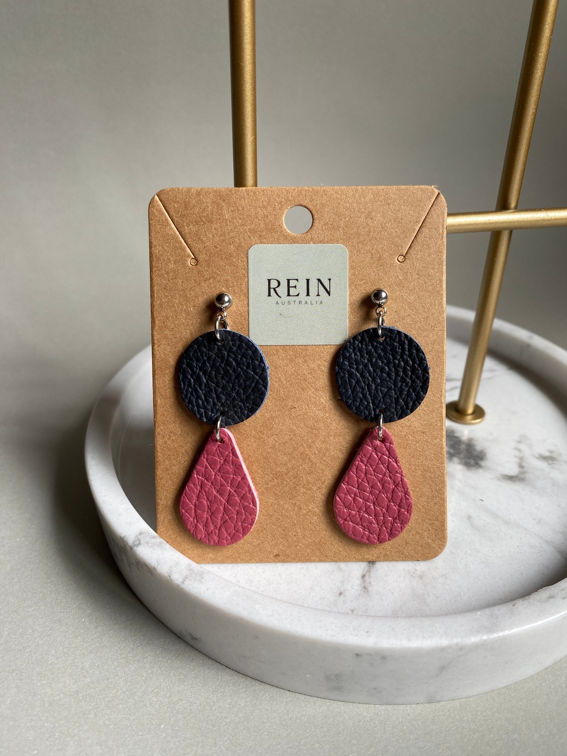 Pair of leather earrings which have a navy circle with a dark pink teardrop suspended below, on a REIN branded card against a neutral background