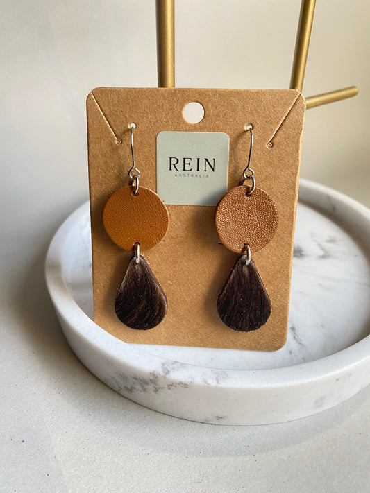 Pair of leather earrings - tan circle with a dark brown hair-on teardrop suspended below, with a silver hook against a marble, grey and gold background with REIN branding.