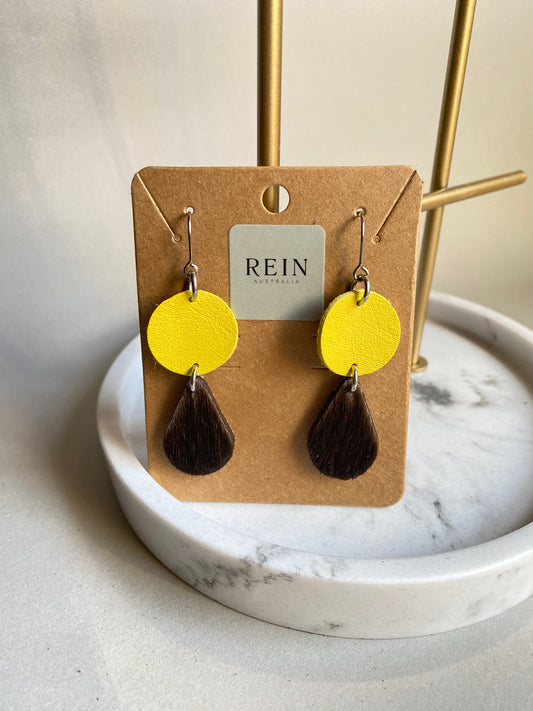 Pair of leather earrings - yellow circle with a dark brown hair-on hide teardrop suspended below, with a silver hook against a marble, grey and gold background with REIN branding