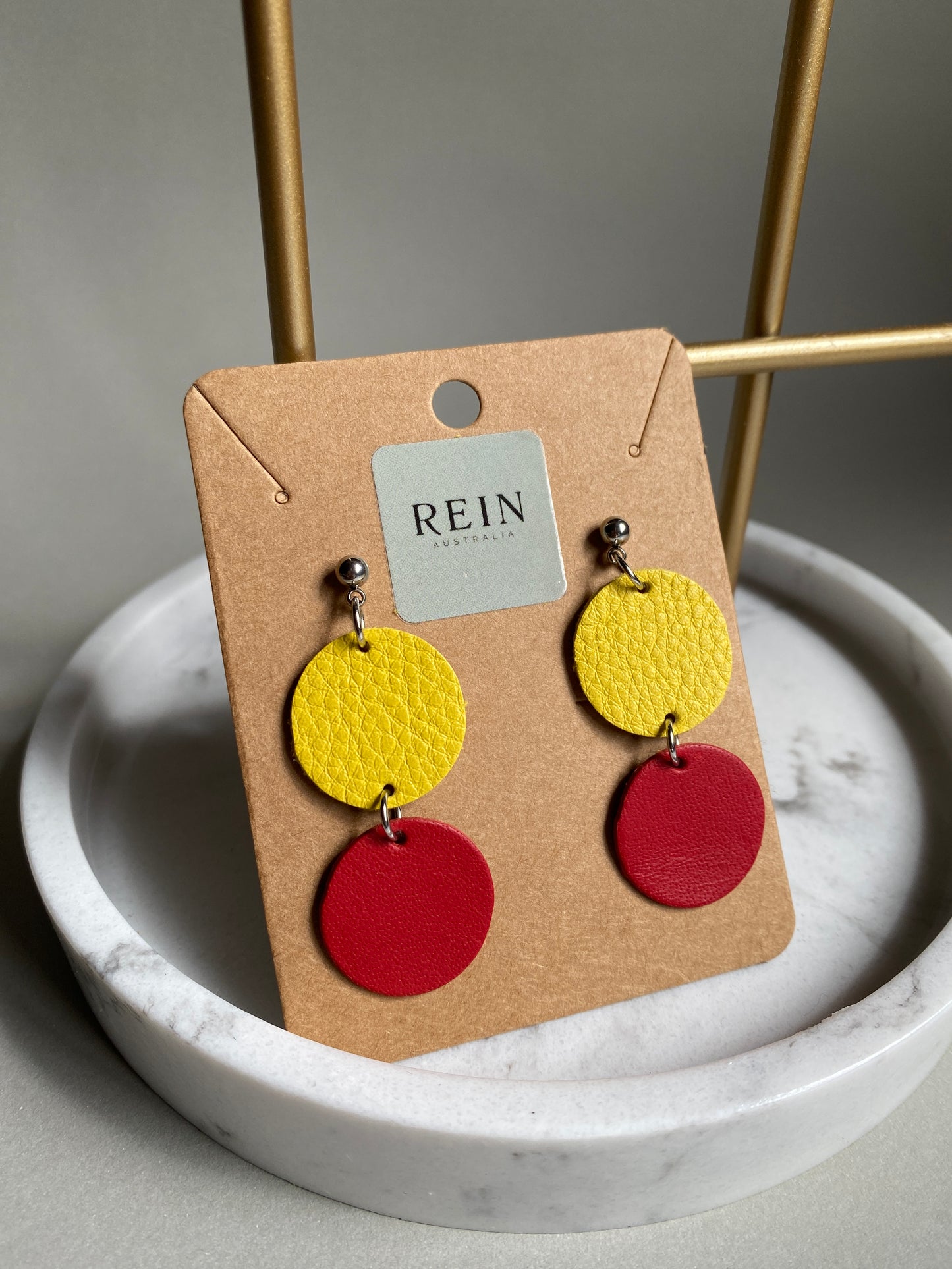 Pair of leather earrings which have a yellow circle with a red circle suspended below, on a REIN branded card against a neutral background