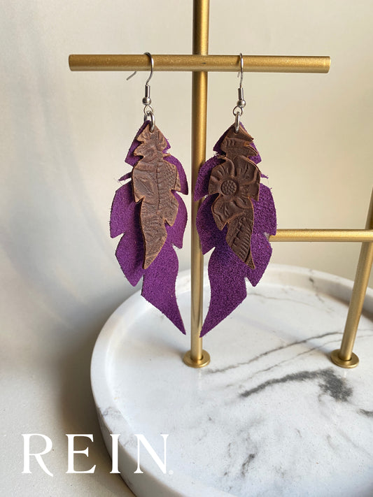 Pair of leather earrings - dark brown embossed leather feather layered over a larger purple suede feather with a silver hook against a marble, grey and gold background with REIN branding