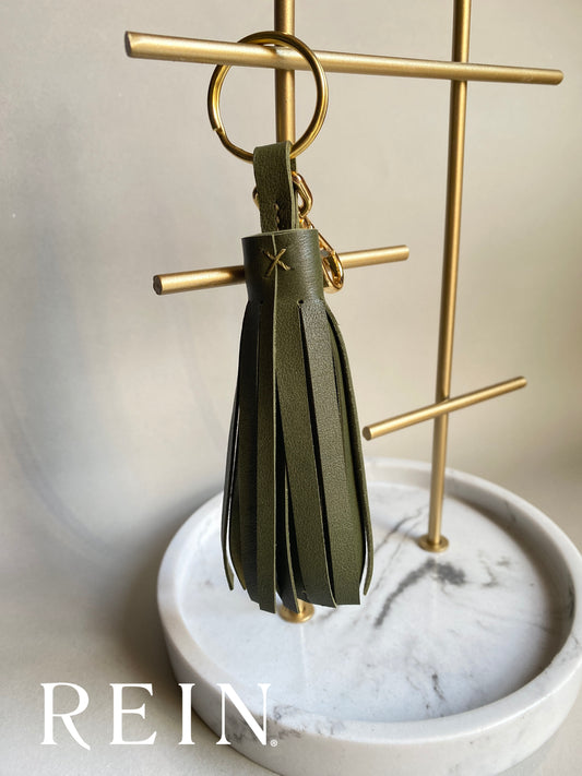 Olive green leather tassel keychain on a gold stand with 'REIN' branding.