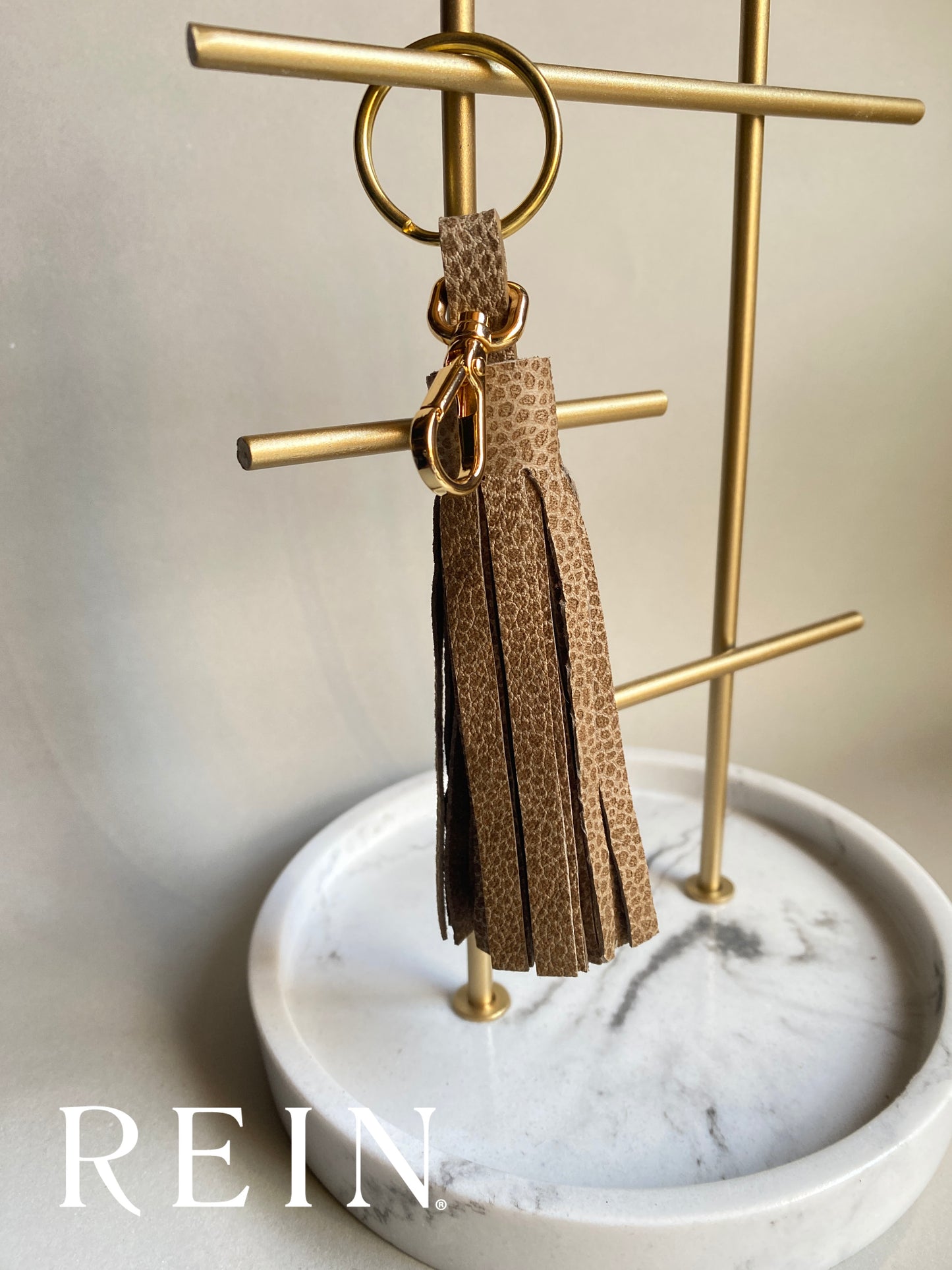 Taupe speckle patterned tassel keychain with a gold snap on a gold stand with 'REIN' branding.