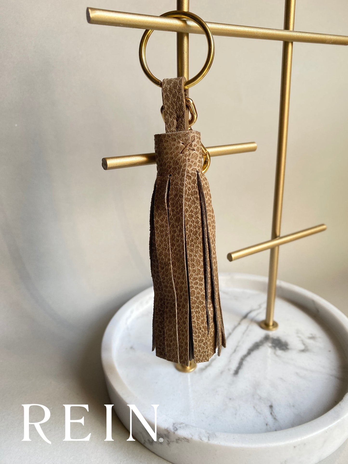 Taupe speckle patterned tassel keychain on a gold stand with 'REIN' branding.