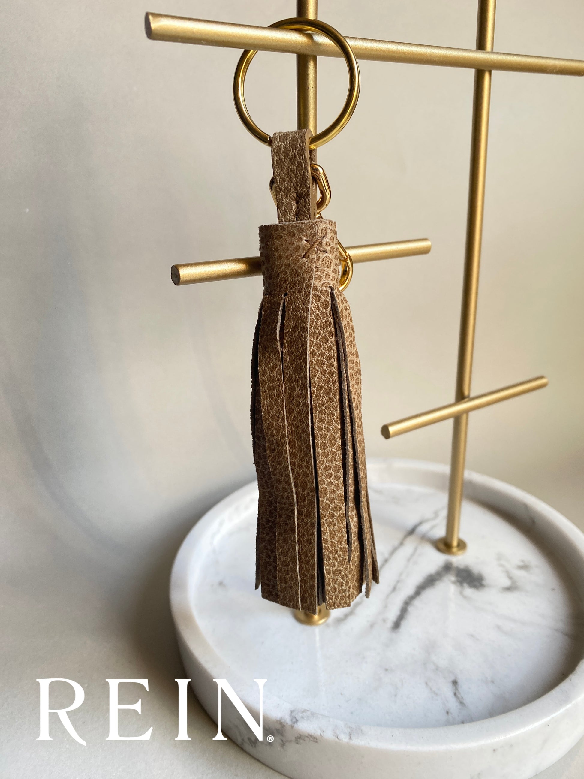 Taupe speckle patterned tassel keychain on a gold stand with 'REIN' branding.