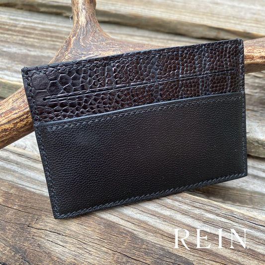 Black leather cardholder with ostrich texture on a wooden surface, featuring the brand 'REIN'.